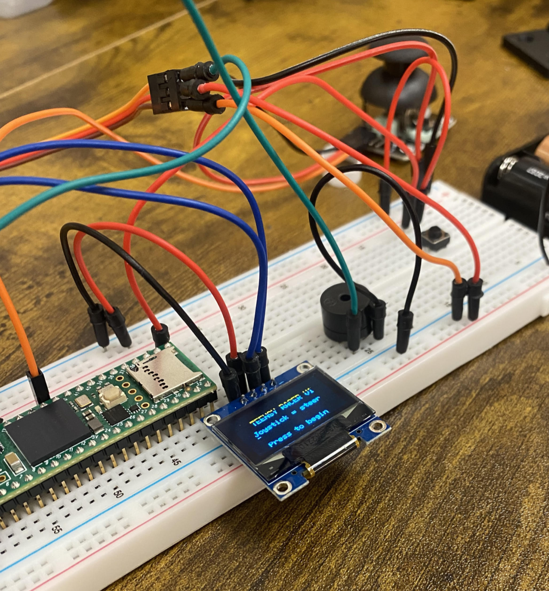 Teensy microcontroller board connected to a breadboard with various multicolored jumper wires, a small LCD screen displaying 'TEMPERATURE: 25.0 C', and a black speaker on a wooden table.